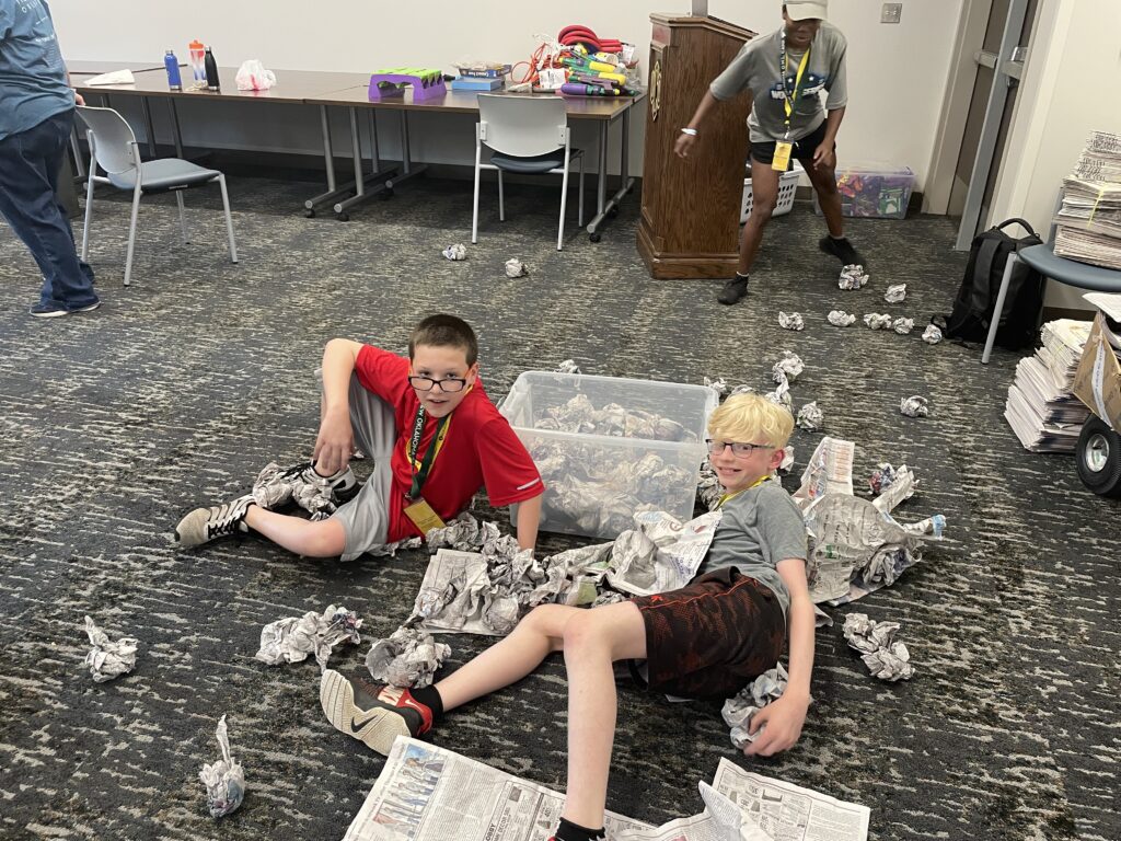Two campers pose for a photo during the Newspaper night. Crumpled up newspapers are all around them.