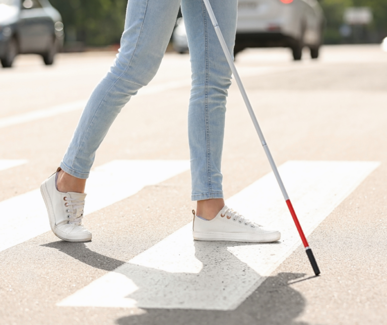 A woman with a white cane is seen crossing at cross walk.
