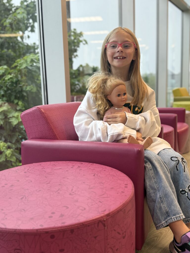 Bristol, an 8-year-old girl with pink glasses, sits in a pink chair holding her doll and smiling at the camera.