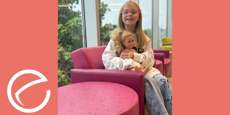 Bristol, an 8-year-old girl with pink glasses, sits in a pink chair holding a doll and smiling at the camera.