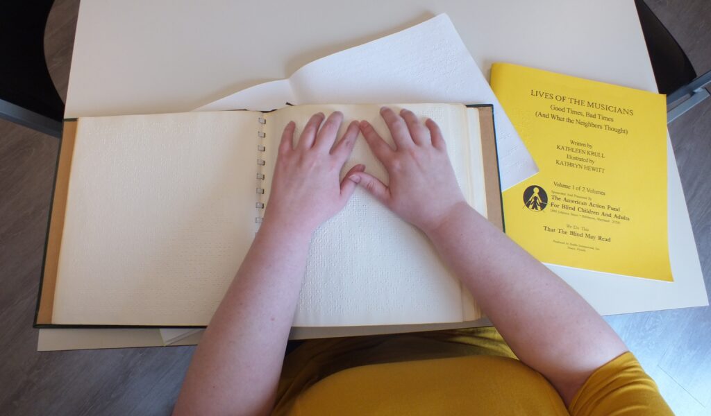 Hands poised to read a braille book, with a second braille volume titled "Lives of the Musicians" nearby.