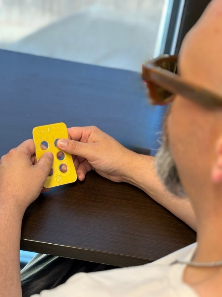 A man wearing sunglasses holds a yellow braille learning card with raised dots, examining it with his fingers.