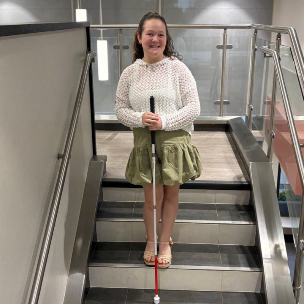 Bella holding her white cane, wearing a cute outfit, smiling and standing on the stairs at NewView.