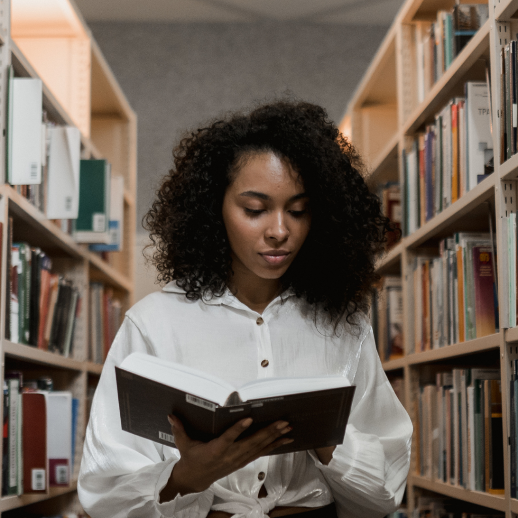 young woman standing between two full bookshelves reading a large print book.